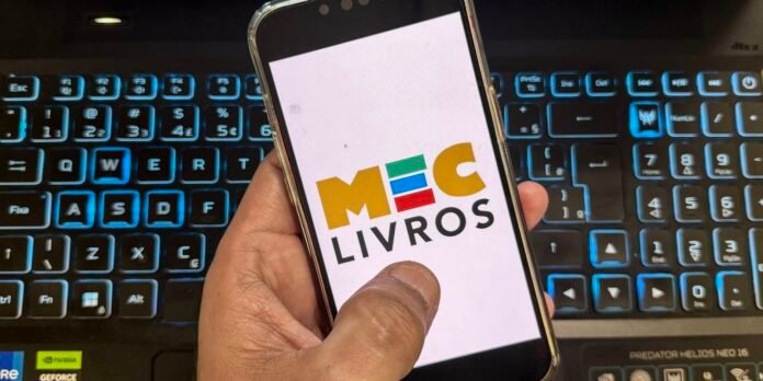Hand holding a smartphone displaying the colorful 'MOC LIVROS' logo over a blue-lit laptop keyboard in the background.
