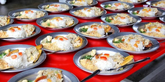 Rows of metal plates each holding white rice with vegetables and crispy toppings on a red table, ready to serve at a buffet.