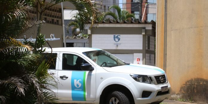 White pickup truck with Sabesp logo parked beside a yellow wall, tropical plants framing the scene.