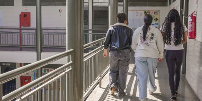 Three students walk down a sunlit school corridor with metal railings and bulletin boards on the walls.