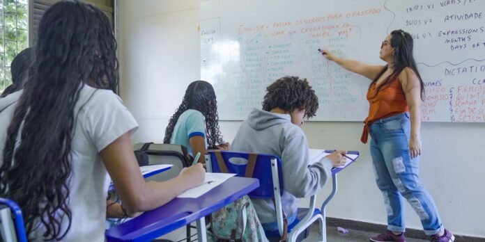 Teacher pointing at a whiteboard filled with English verbs while students take notes in a bright classroom.