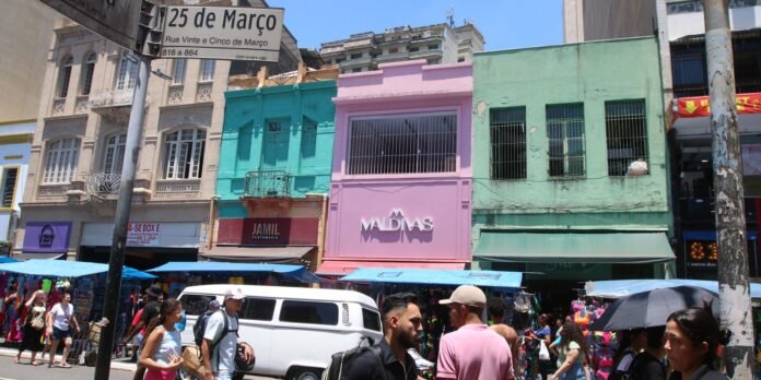 Busy street market in front of pastel-colored buildings, with blue awnings and shoppers beside a white van.