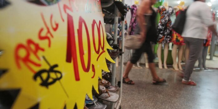 Shoes on display in a store with a bright yellow sale sign in the foreground and shoppers walking by in the background.