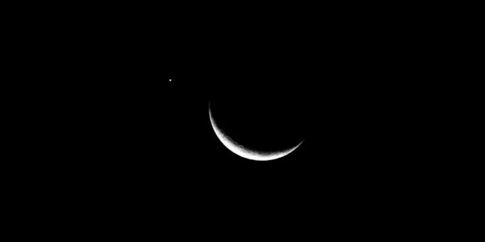 Slim crescent Moon glowing white against a black night sky with a bright dot nearby (likely a star or planet)