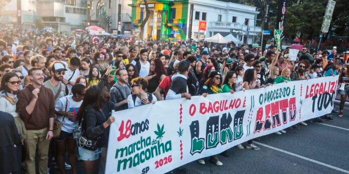 Large crowd of people marching in a city street, holding a long banner with green and red text that reads '18 anos marcha da maconha SP 2026' and has marijuana leaf graphics; urban background with storefronts