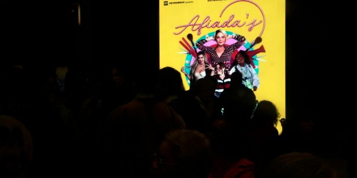 Bright yellow promotional poster featuring a group of women with makeup brushes, seen behind a crowd in a dark room.