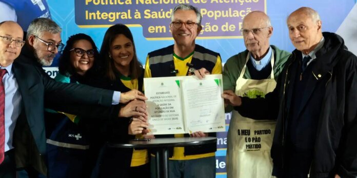 Seven adults stand together around a table, holding an open certificate as they smile for the photo, with a blue banner in Portuguese in the background reading about national health policy for the homeless.