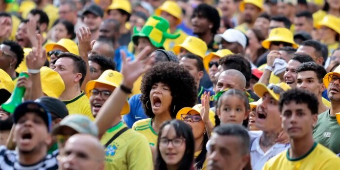 Crowd of Brazil fans in yellow shirts and hats cheering at a stadium, with a woman with curly hair shouting in the foreground.