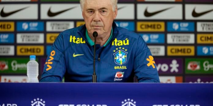 Brazil national team coach at a press conference, speaking into a microphone, wearing a blue jacket with Brazil crest and sponsor logos.