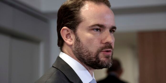 Side profile of a bearded man in a dark suit and light blue tie, speaking in an indoor setting.