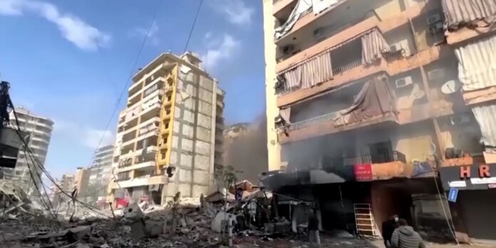 Rubble-strewn urban street with collapsed buildings, tangled wires, and smoke in the air; a few people observe the debris.