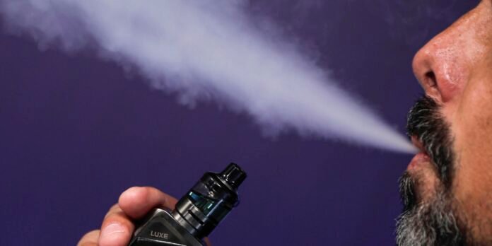 Close-up of a man with a beard exhaling vapor from a Luxe vape pen held in his hand against a dark purple background.