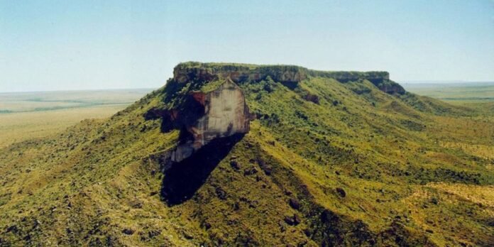 Flat-topped mesa with green slopes rising from a vast, sunlit grassland under a clear blue sky.