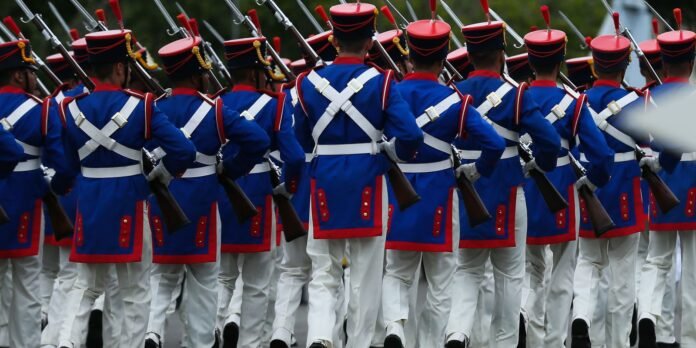 Rows of soldiers in blue uniforms with red trim march in a parade, rifles raised in formation.