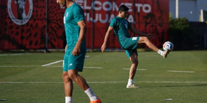 Two soccer players in teal uniforms train on a green field; one raises a leg to kick the ball while the other stands nearby, with a red banner backdrop.