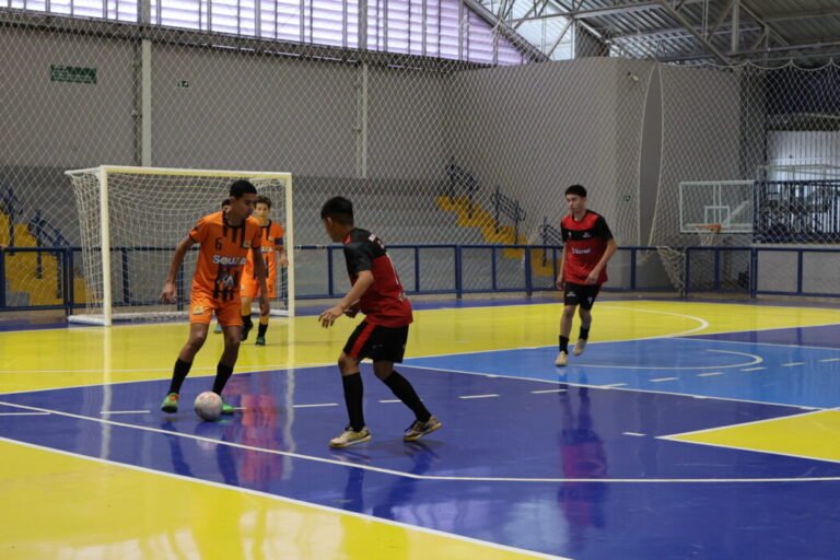 Indoor futsal match: orange and red team players contest the ball on a blue-and-yellow court with a goal in the background.