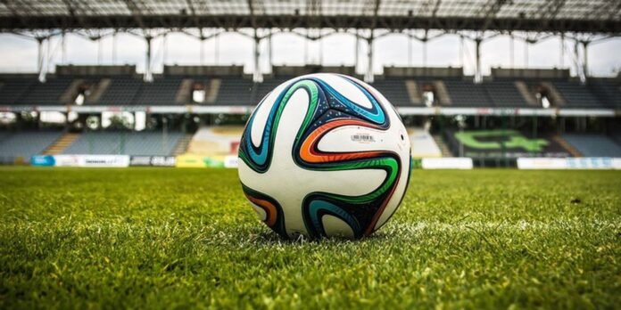 Soccer ball resting on a green grass field inside an empty stadium with blurred seating in the background.
