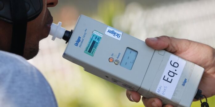 A man blows into a digital breathalyzer held by another person, measuring blood alcohol content outdoors.