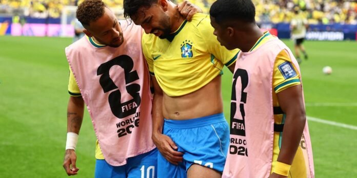 Three Brazil teammates in yellow kits with pink bibs share a moment on a green field, one player helping another off the pitch edge.