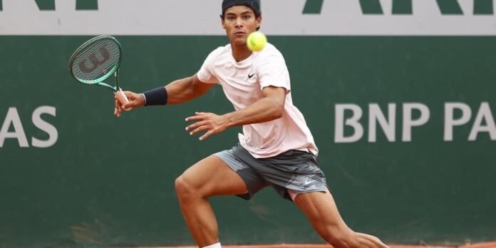 Male tennis player in a white shirt and gray shorts lunges to return a yellow ball with a racket on a green court, left arm extended.
