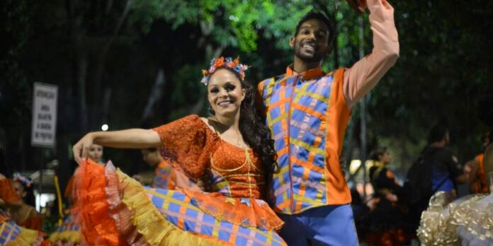 Couple dancing at night in bright, folkloric outfits; the woman wears an orange dress with a flower crown, and the man wears a multicolored vest and orange shirt as they pose mid-dance.