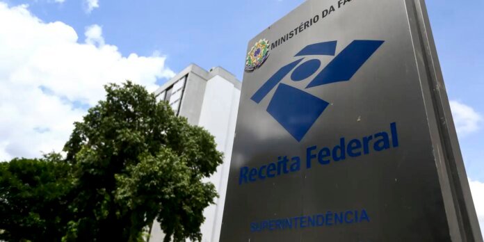 Metal sign reading 'Receita Federal' with blue logo in front of a government building and trees under a blue sky; Brazil’s federal revenue office.