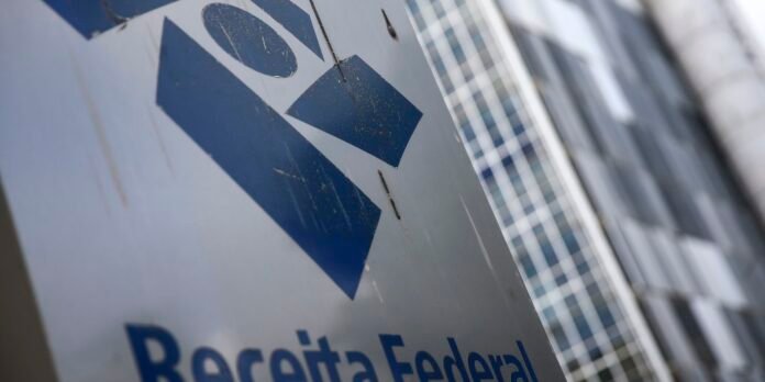 Close-up of a weathered metal sign with a blue geometric logo, and partial text, set against a modern glass building.