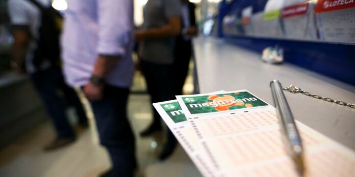 Two Mega-Sena lottery tickets lay on a counter next to a silver pen, with blurred customers standing in line in the background.