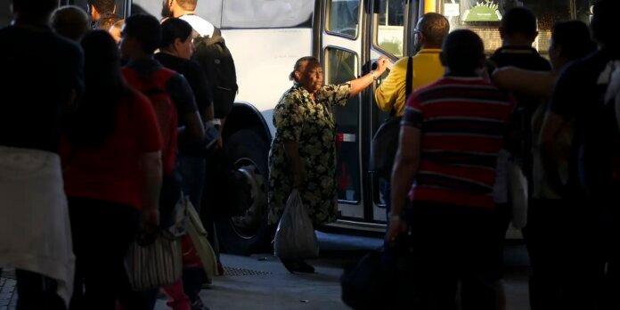 Older woman in a floral dress exits a bus, carrying a plastic bag, as a crowd waits nearby on a city street.