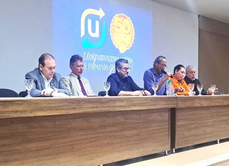 Six people seated at a long wooden panel desk during a conference, with a blue backdrop featuring a 50th‑anniversary emblem behind them.
