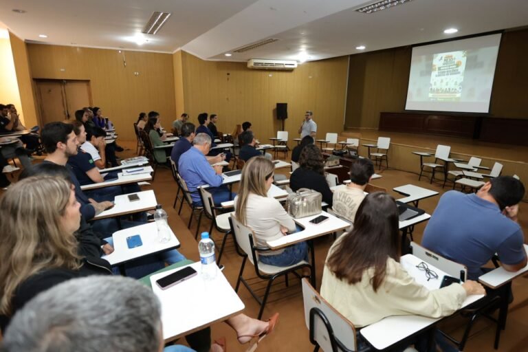 Speaker presenting to a classroom audience in a lecture hall with students taking notes and laptops open, projector screen on the front wall showing a poster at right.