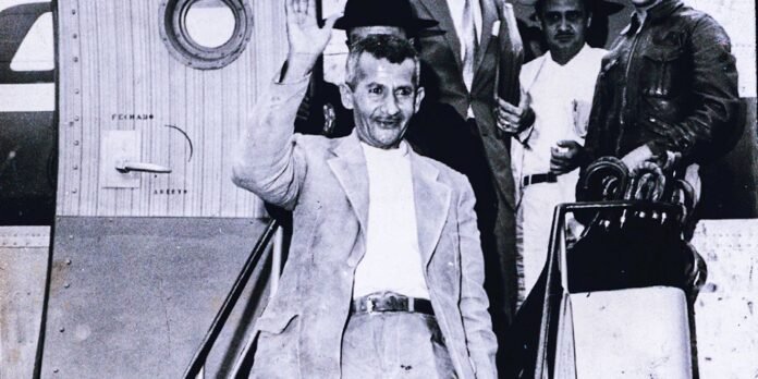 Man in a light blazer and white shirt waves from airplane stairs while a group of men stands behind him beside the door of the plane.