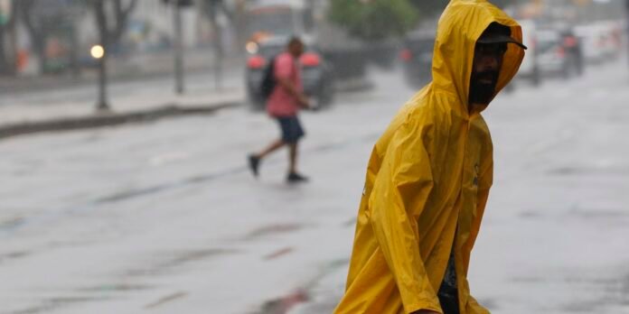 Person in a bright yellow raincoat walking through heavy rain on a city street, with cars and another pedestrian blurred in the background.