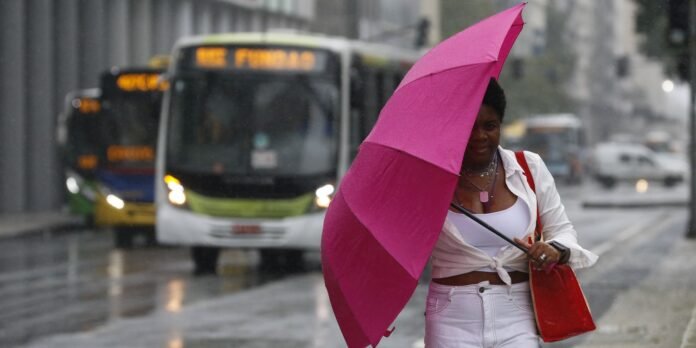 Woman walking in the rain with a bright pink umbrella, wearing white clothing and carrying a red bag on a wet city street with buses in the background.