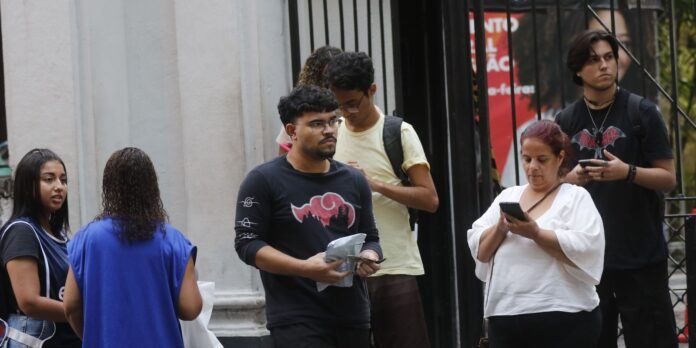 Group of diverse people standing outside a building, some looking at phones and bags in hand.