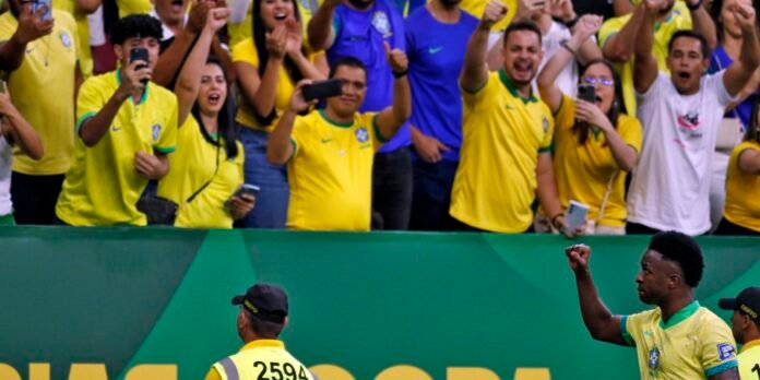 Brazilian soccer player raises a clenched fist as fans in yellow cheer and film from behind the barrier