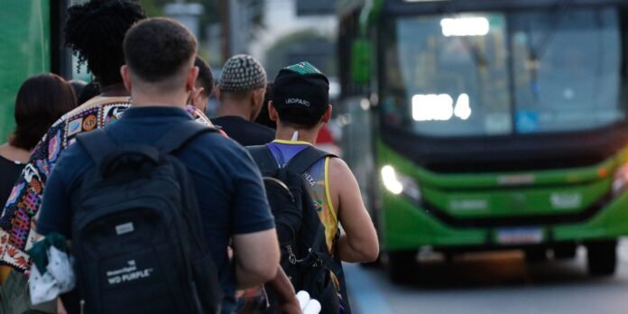People line up with backpacks in front of a green bus on a city street, waiting to board.
