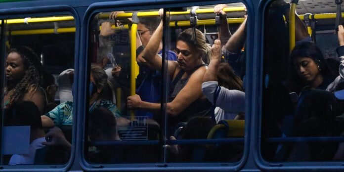 Crowded city bus interior with passengers standing and gripping yellow overhead poles near windows/doors.
