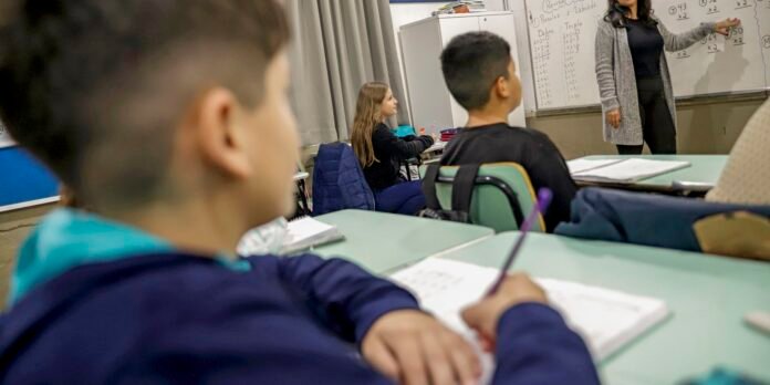 Teacher explains math on a whiteboard at the front of a classroom as students listen and take notes.