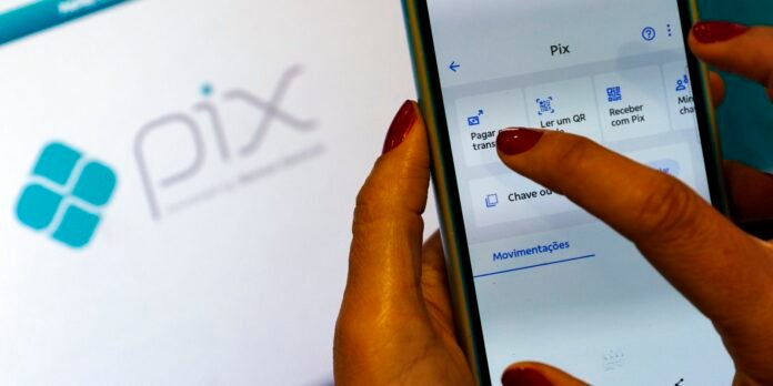 Close-up of hands with red nails using a smartphone to access the Pix app, with a blurred Pix logo in the background.
