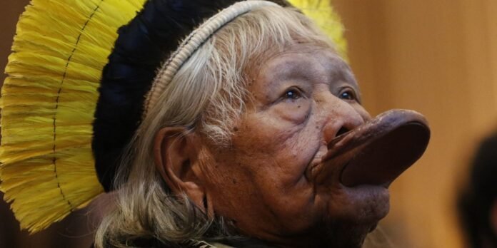 Older person wearing a yellow feather headdress and a large pig-nose prosthetic, looking upward to the right.