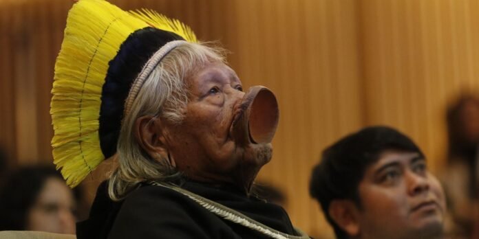 Elderly Indigenous person wearing a bright yellow feather headdress and a large ceremonial nose plate, indoors with audience in the background.