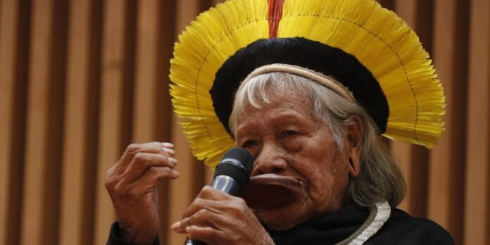 Elderly Indigenous woman wearing a bright yellow feather headdress and speaking into a handheld microphone at a podium or stage.
