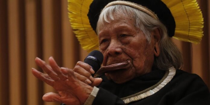 Indigenous elder in a black robe and yellow feather headpiece speaks into a microphone, hands raised in gesture of speaking