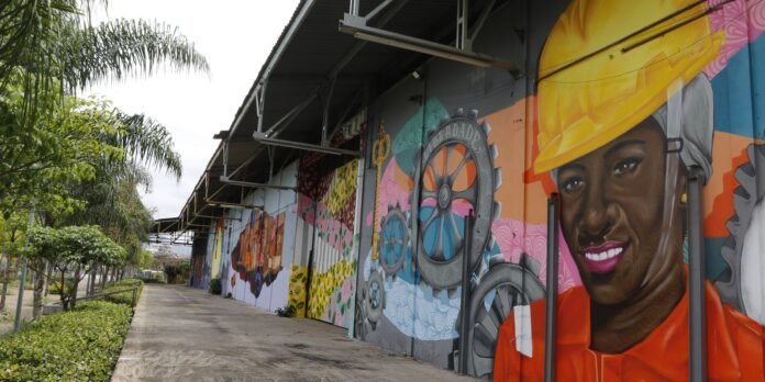 Mural on an industrial building showing a smiling woman in an orange work outfit and yellow hard hat, with gears and colorful patterns on the wall.