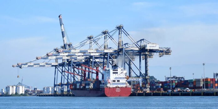 Red cargo ship named HARIS VALLETTA at a busy container port with tall blue gantry cranes and stacked shipping containers in the background, calm water in the foreground.