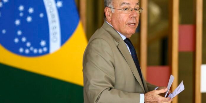 Man in a beige suit and glasses speaks, holding papers, with a large Brazilian flag in the background.