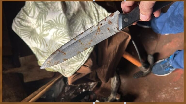 Close-up of a rust-stained kitchen knife blade held by a gloved hand in a workshop setting, showing brown rust spots along the blade.