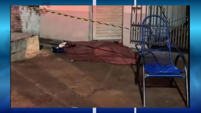 Body covered by a brown tarp on a sidewalk, yellow caution tape across the scene, with a blue metal chair in the foreground and a gated area nearby.