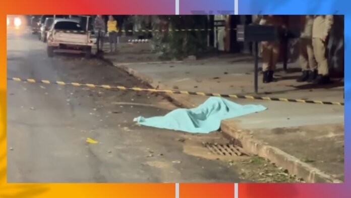 Body covered by a light blue sheet lying on a sidewalk and street, with yellow police tape and officers in the background at a nighttime crime scene.